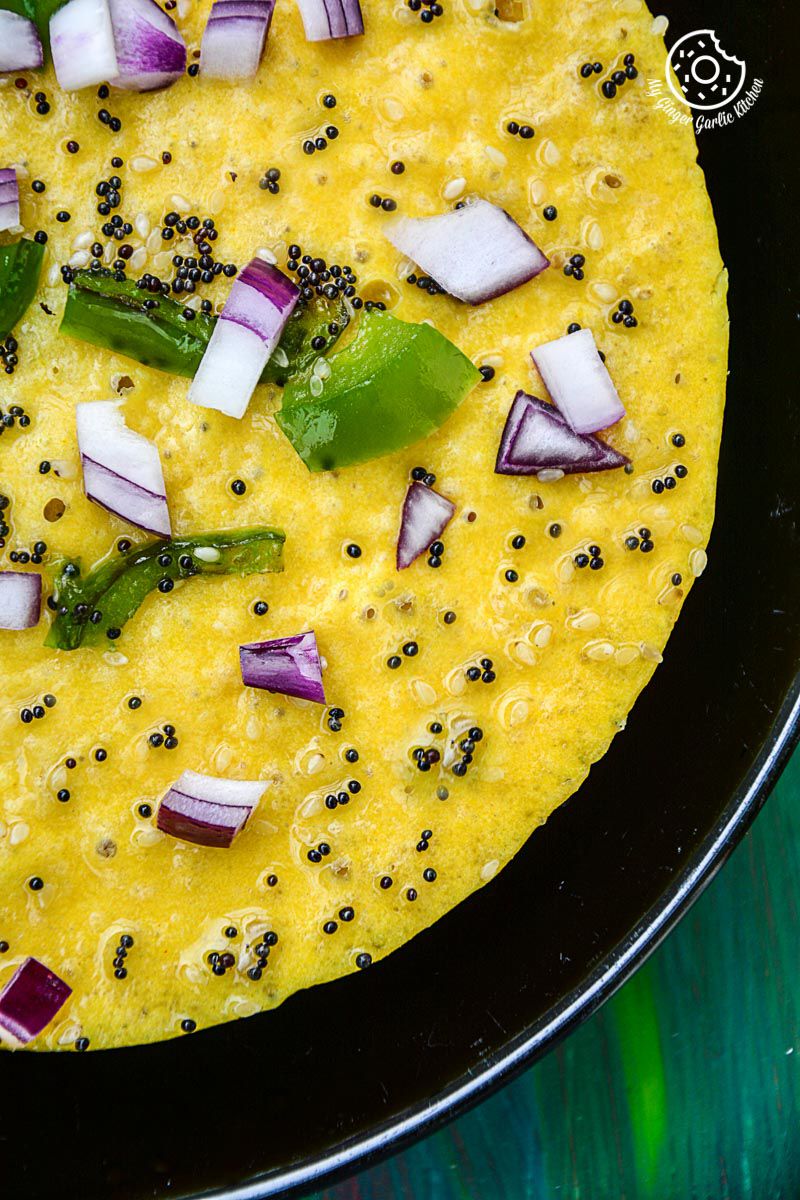 a black pan with a gujarati khaman dhokla topped with tempering, onions and peppers