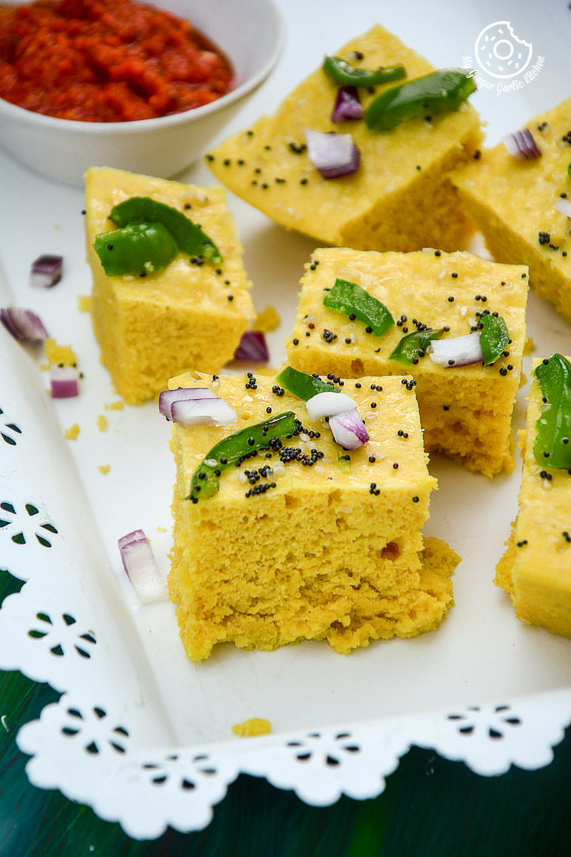 a plate of gujarati khaman dhokla pieces with a bowl of red chutney