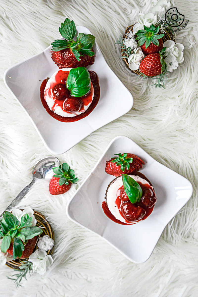 two plates with two small white plates with no bake mini strawberry cheesecake and strawberry compote on them and a cup of strawberries on a white furnish