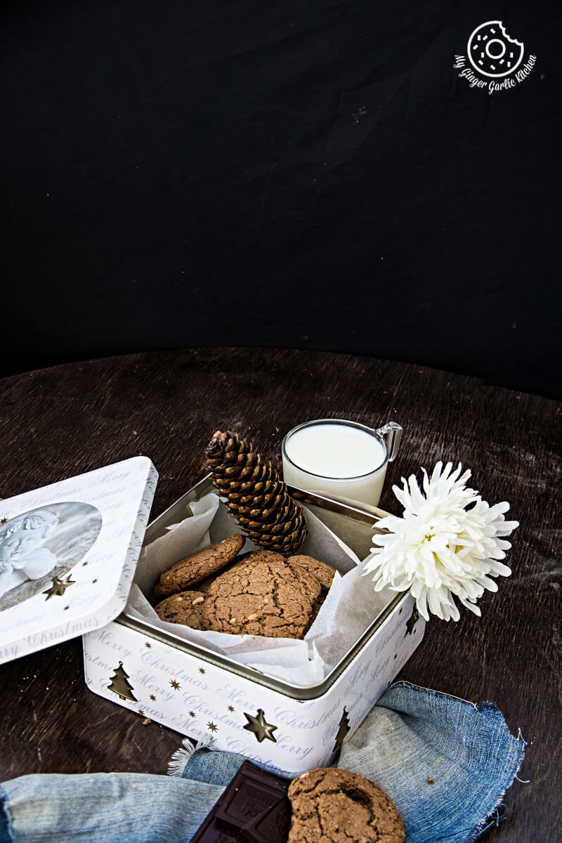 a tin with oatmeal peanut butter chocolate cookies and a glass of milk on a table