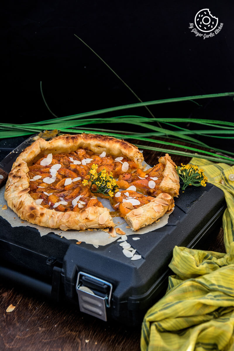 a puff pastry apricot galette with almond topping on a black case with a bunch of vegetables