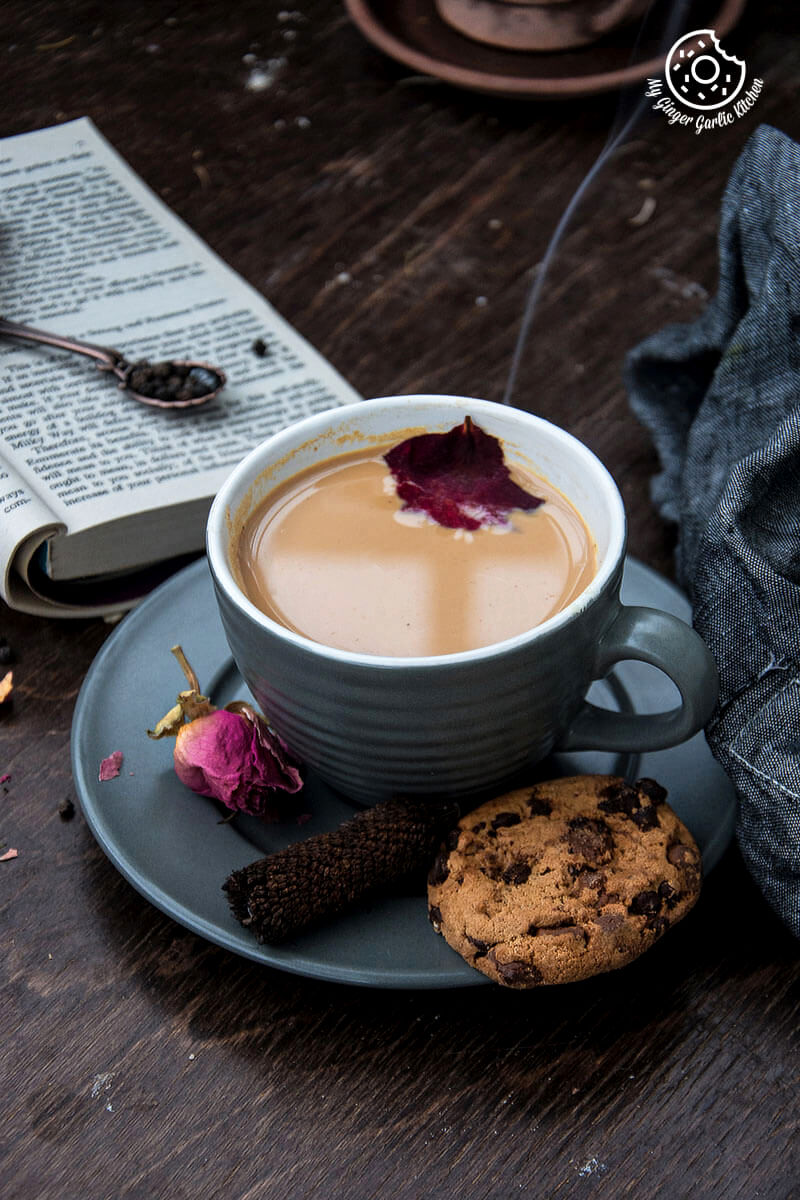 a cup of rose masala chai and a chocolate chip cookie on a table with a book and table cloth