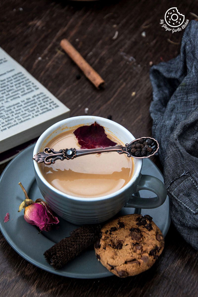 a cup of rose masala chai and a chocolate chip cookie on a table with a book and table cloth