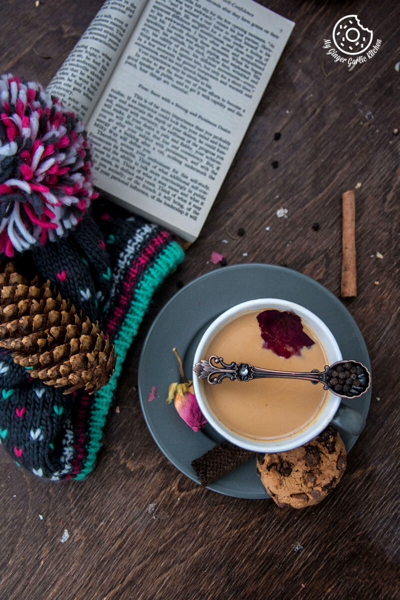 a cup of rose masala chai and a chocolate chip cookie on a table with a book and a hat