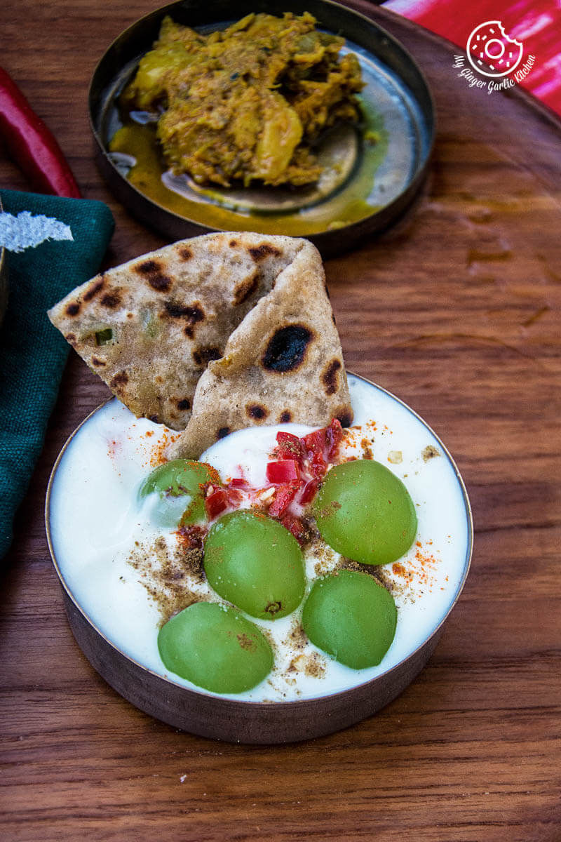a bowl of yogurt with grapes and a piece of cabbage peas stuffed paratha