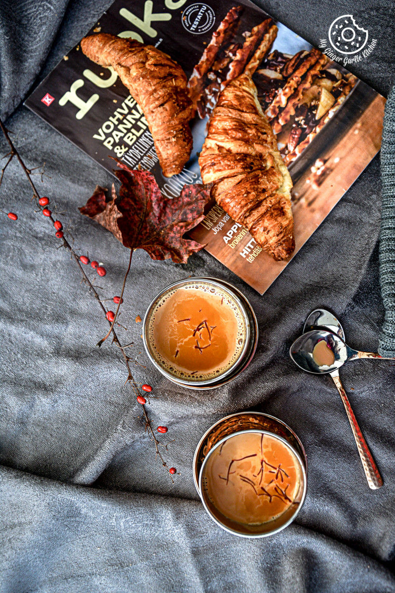 there is a book and two metal glasses of spiced kesar chai topped with saffron strands