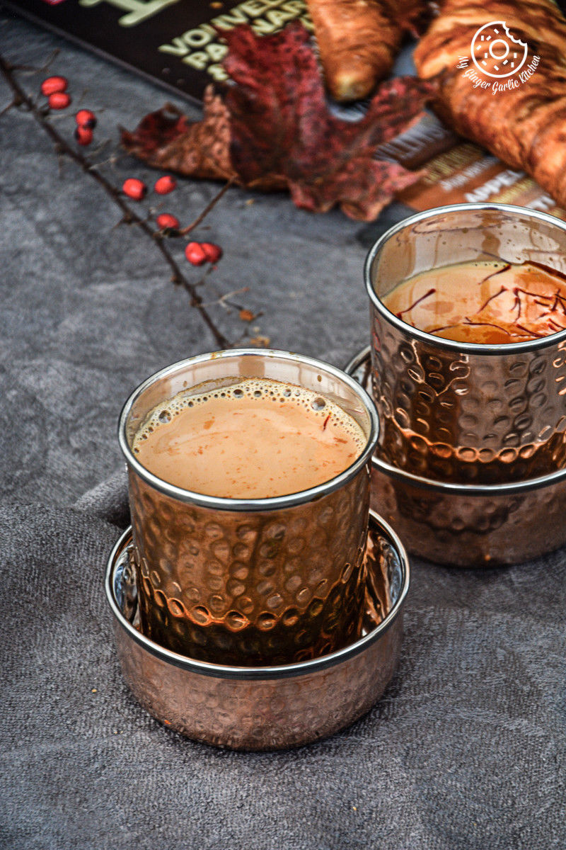 there are two cups of spiced kesar chai topped with saffron strands on a table with a book