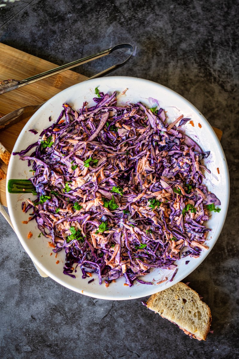 A bowl of vibrant red cabbage coleslaw mixed with shredded carrots and garnished with fresh cilantro, placed on a wooden cutting board with a serving tong nearby.