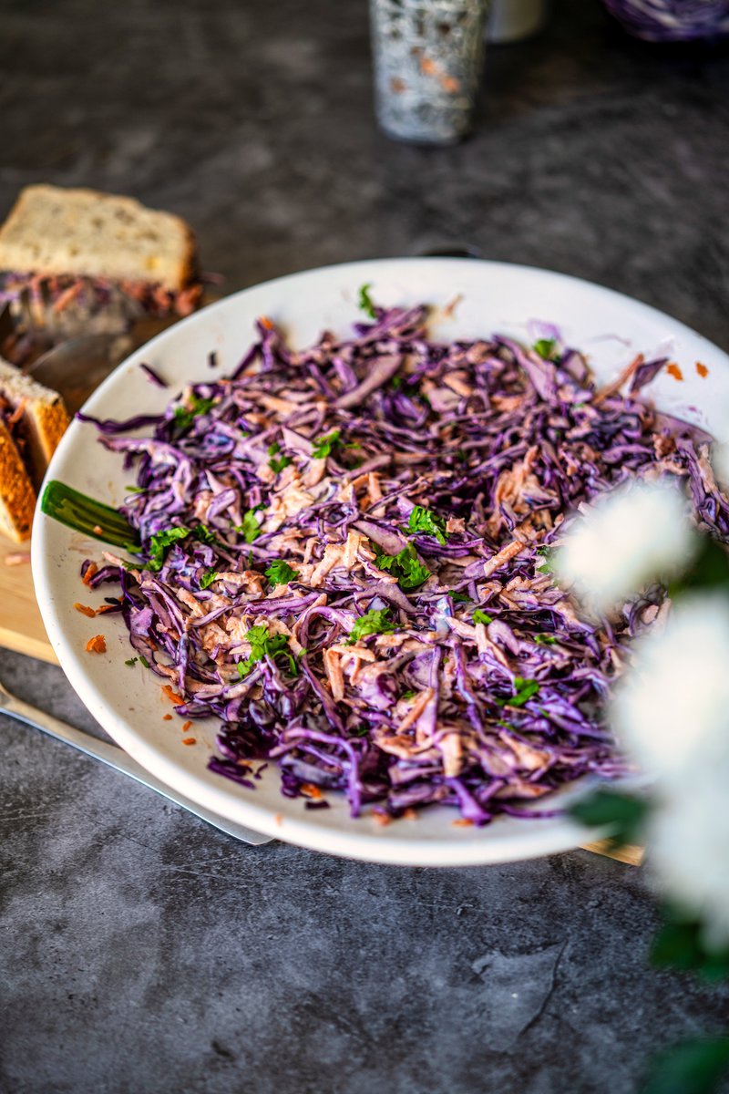 A close-up of red cabbage coleslaw in a white bowl, showcasing the purple cabbage and orange carrots, garnished with fresh herbs, on a gray countertop with a sandwich in the background.