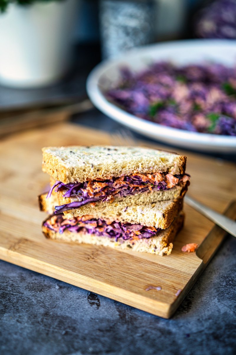  A sandwich made with whole wheat bread, stuffed with creamy red cabbage coleslaw, sitting on a wooden board, with the coleslaw bowl in the background.