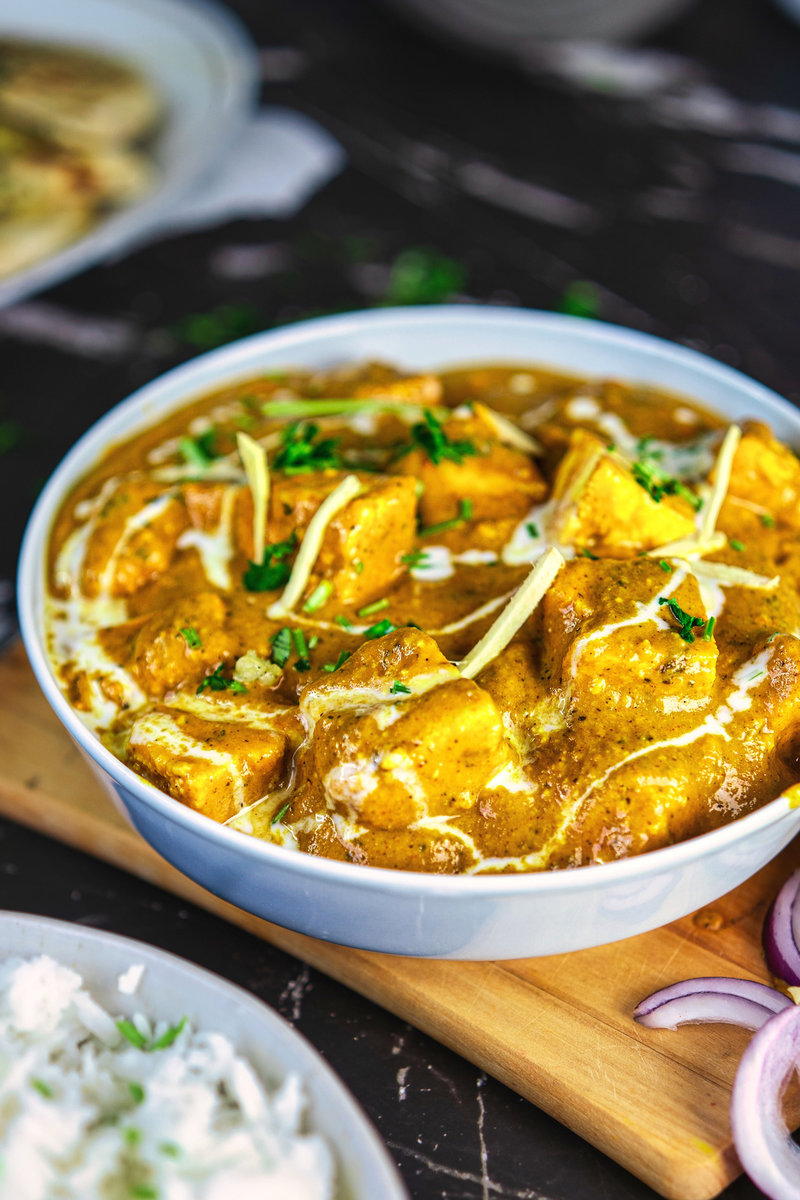 A close-up of paneer butter masala garnished with ginger juliennes and fresh cilantro in a white bowl, with blurred naan and rice in the background.