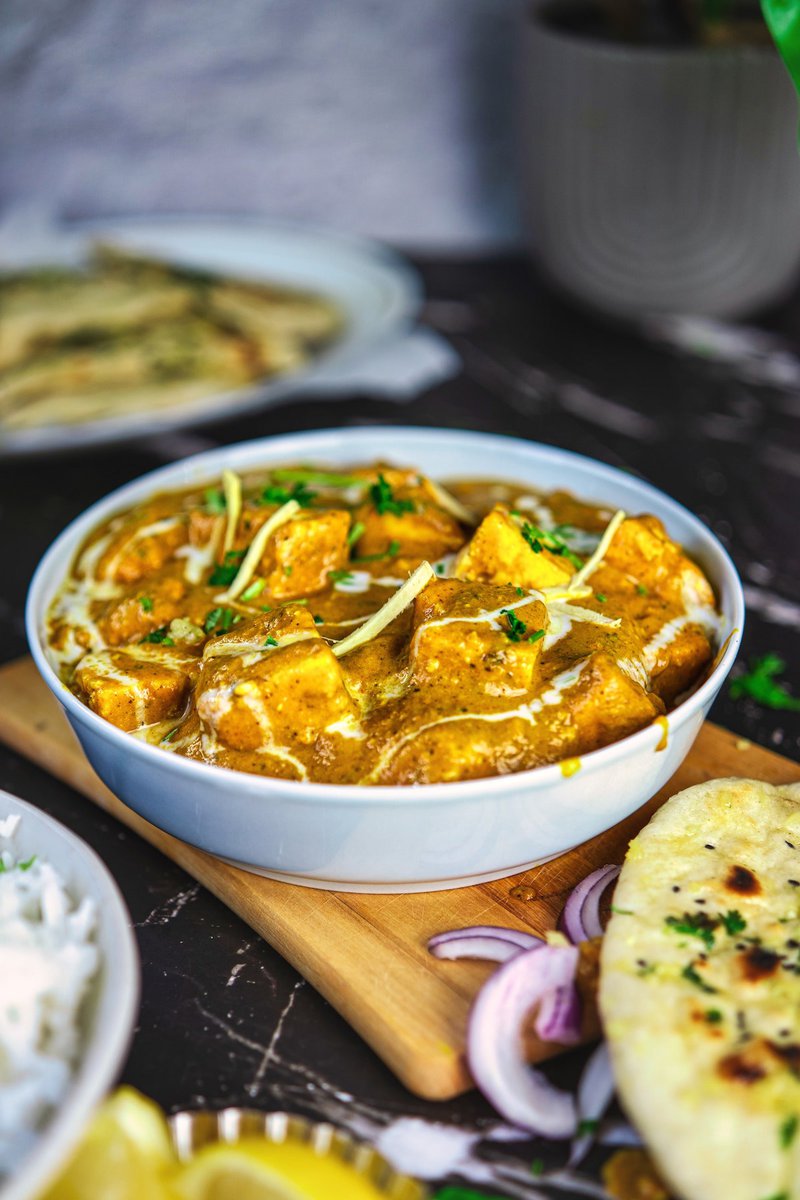 Paneer butter masala in a white bowl on a wooden board, accompanied by naan and sliced onions, with a hint of rice and raita in the back.