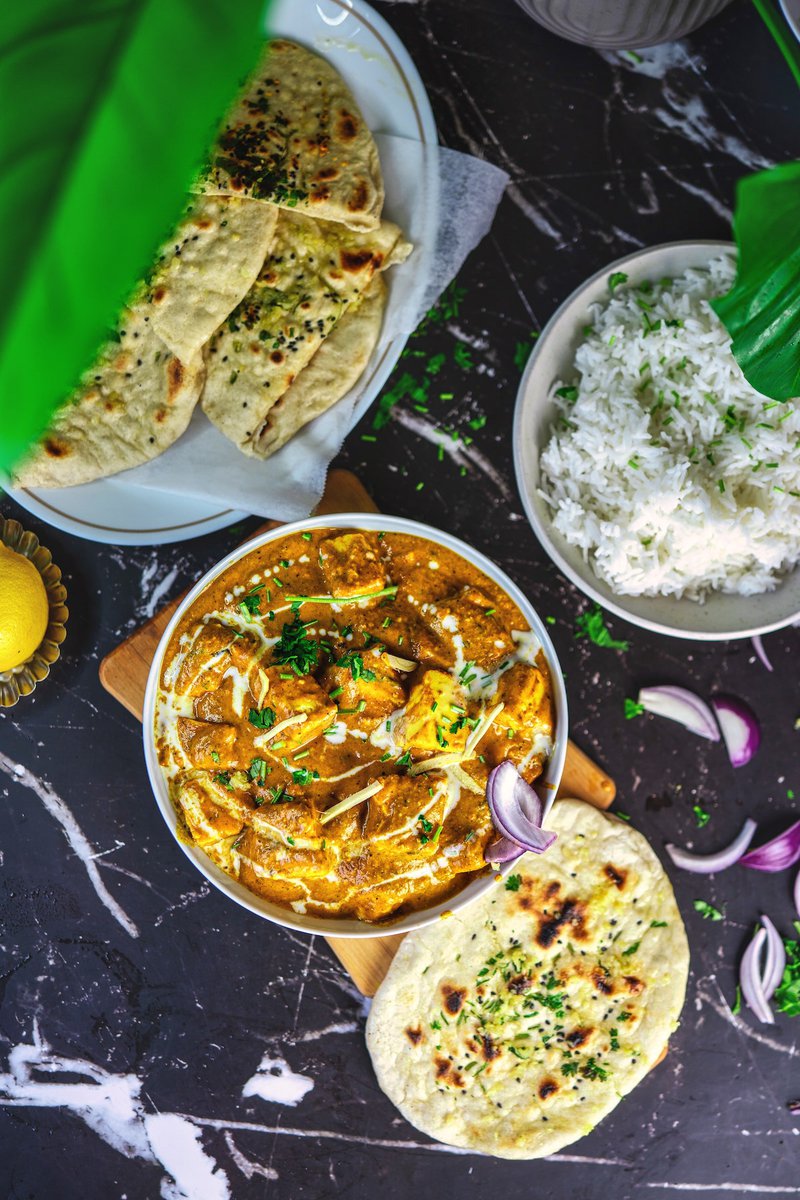 Paneer butter masala served in a white bowl on a dark surface, flanked by naan, rice, and sliced onions, with vibrant greenery in the corner adding a pop of color.