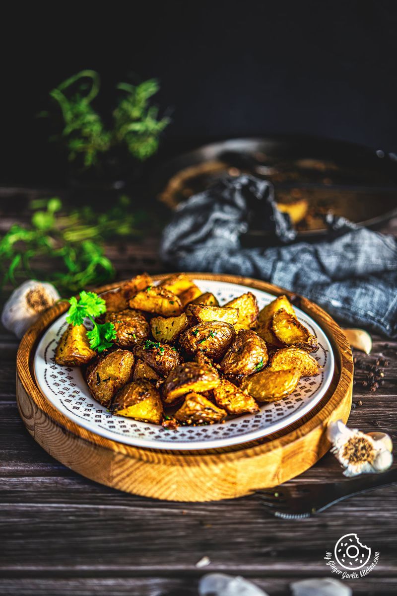 oven roasted potatoes in a white plate with some garlic bulbs and rosemary leaves in the background