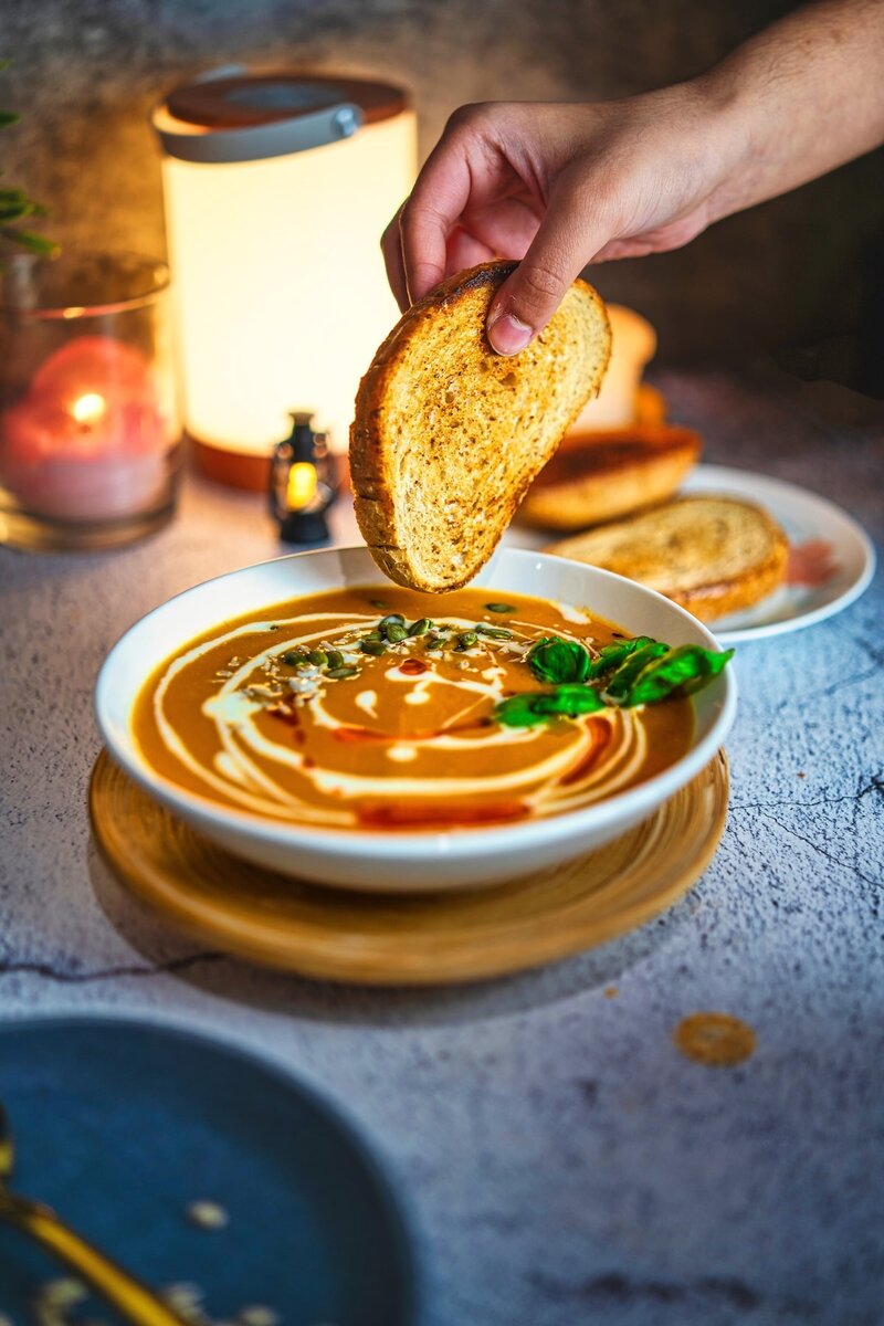 Hand dipping toasted bread into creamy vegan roasted tomato soup topped with basil, seeds, and chili oil