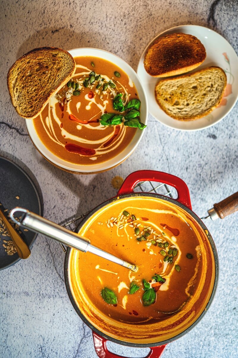 Top view of fresh roasted tomato soup and toasted bread with basil, cream swirl, and crunchy seeds.