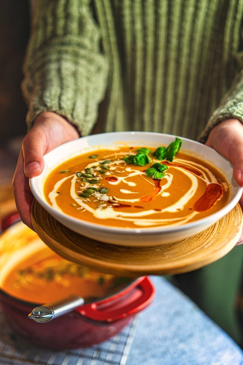 Hands holding a bowl of creamy vegan roasted tomato soup with basil, cream drizzle, and pumpkin seeds.