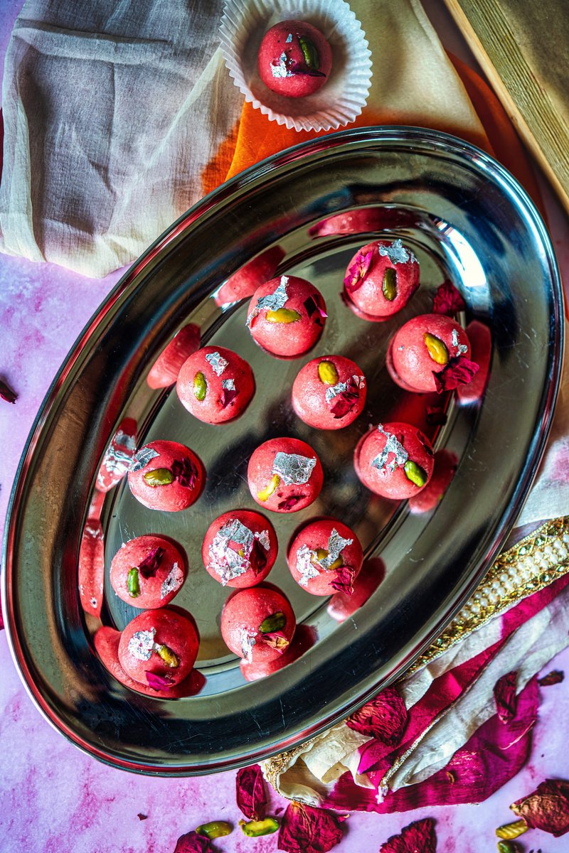 A tray of rose badam ladoos garnished with silver leaf and pistachios, surrounded by rose petals and fabric.