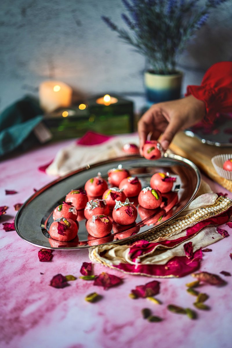 A platter of rose badam ladoos garnished with pistachio and rose petals, placed on a pink table amid candles and decorative elements.