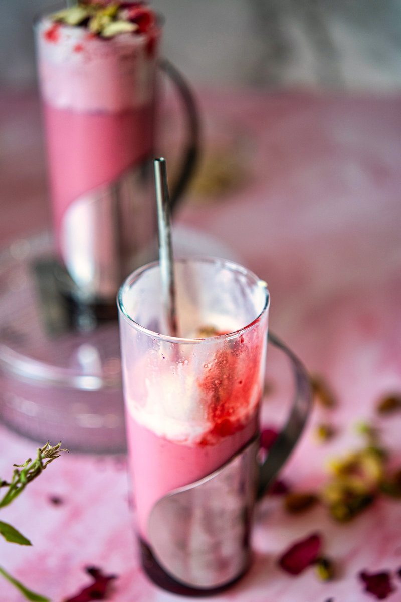 Glass of vibrant pink rose milkshake topped with ice cream, rose syrup, dried rose petals, and chopped pistachios.