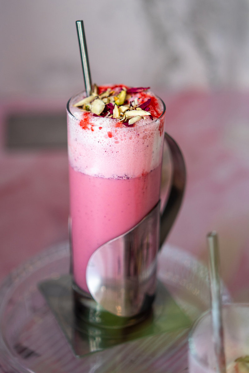 Tall glass of frothy pink rose milkshake garnished with rose syrup, dried rose petals, and chopped pistachios.