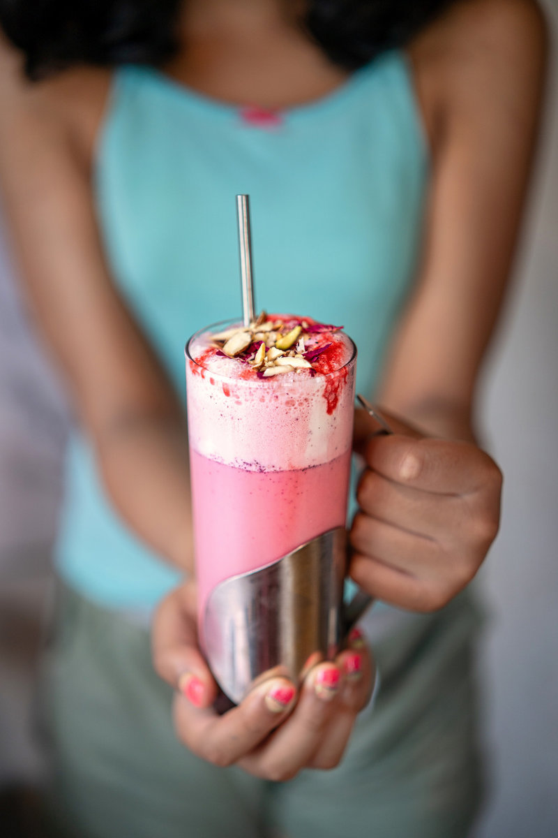 Person holding a tall glass of pink rose milkshake garnished with rose syrup, dried rose petals, and chopped pistachios.