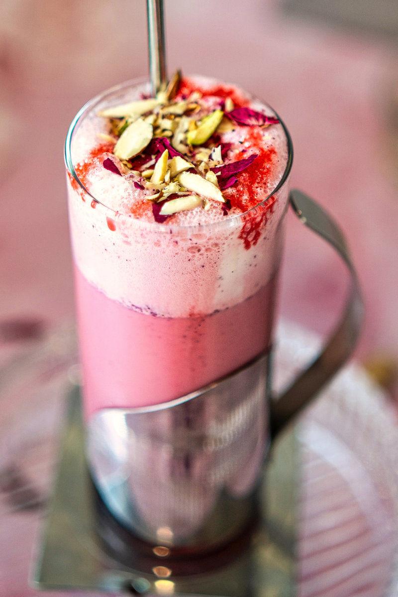 Top-down view of a frothy pink rose milkshake garnished with rose syrup, dried rose petals, and chopped pistachios.