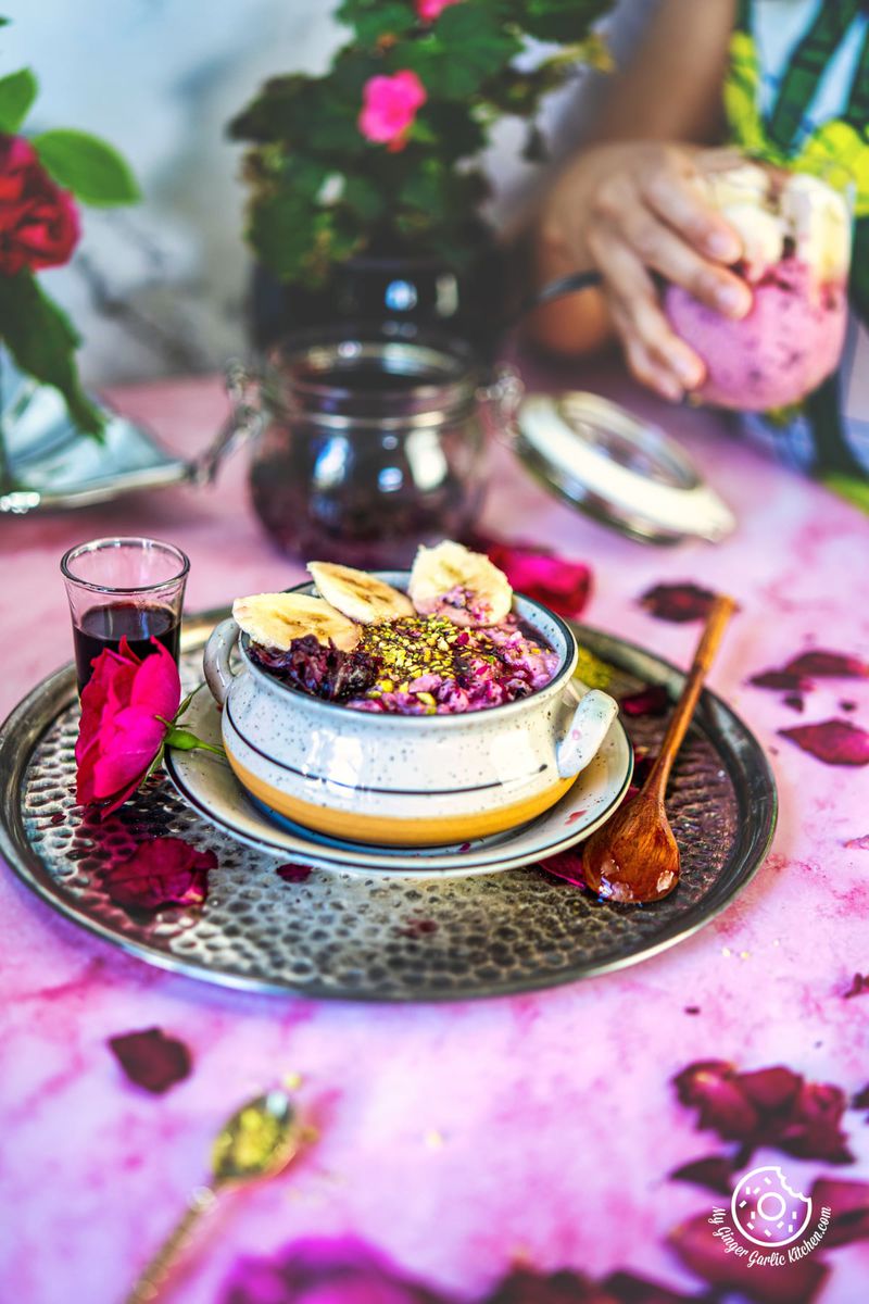 overhead shot of rose overnight oats topped with banana slices, with some red rose petals and rose syrup on the side