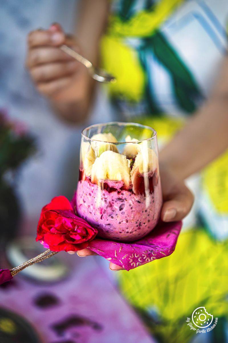 a closeup of a female holding a transparent glass filled with rose overnight oats over a pink cloth and red rose