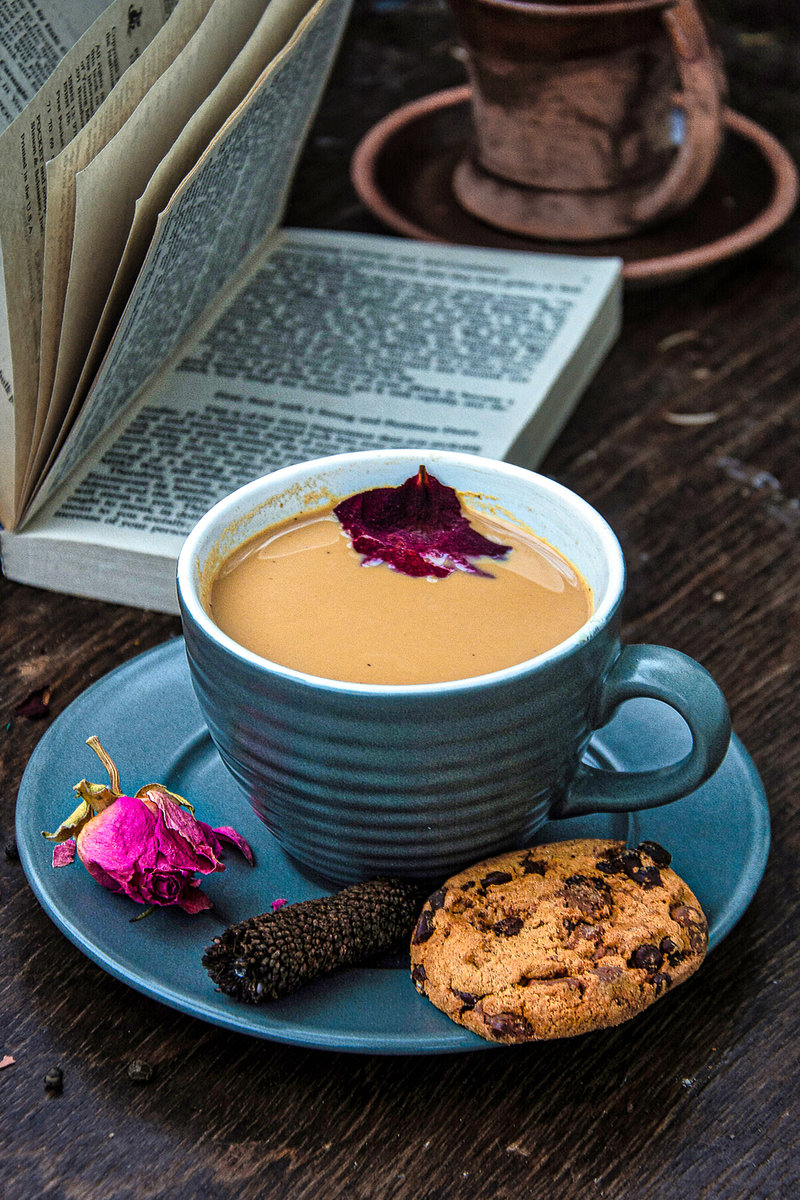 A cozy setting featuring a cup of rose masala chai topped with a dried rose petal, accompanied by a chocolate chip cookie and a dried rose on a teal saucer. A blurred book and a terracotta pot are in the background, set on a rustic wooden surface.