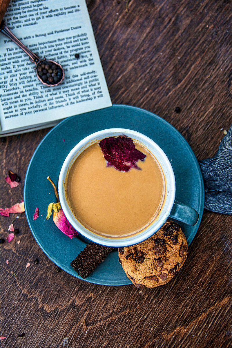 Overhead view of rose masala chai with a vibrant rose petal floating on top, a chocolate chip cookie, and a black spice spoon with chai spices on a teal saucer next to an open book on a wooden table.