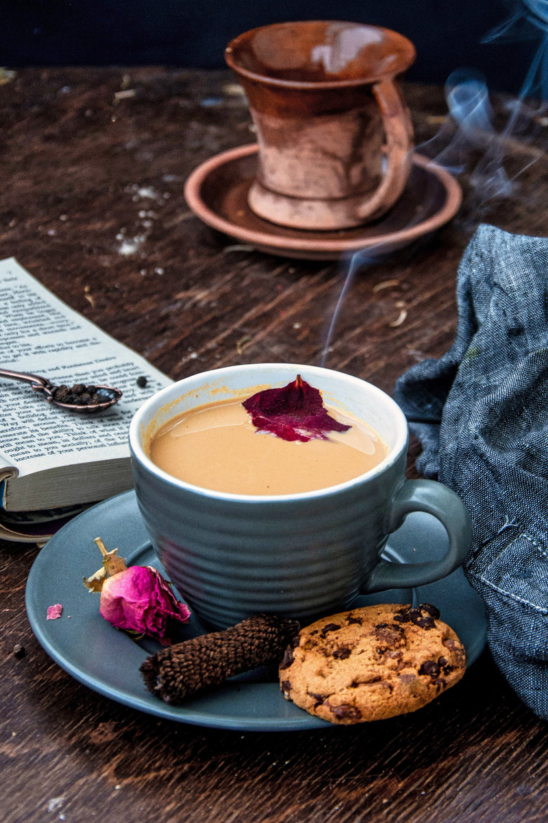 A steaming cup of rose masala chai with a dried rose petal garnish, a chocolate chip cookie, and a spent rose bloom, set beside an open book and a folded cloth napkin, creating a relaxing reading atmosphere.
