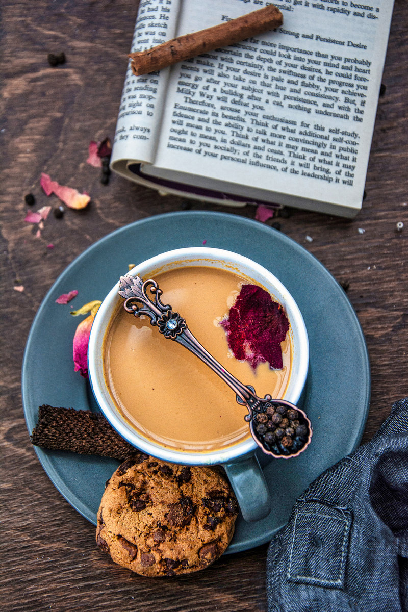 An inviting scene of rose masala chai tea with a single rose petal on the surface, a chocolate chip cookie, and an ornate spoon resting on the saucer, with an open book and loose rose petals scattered around on a wooden table.