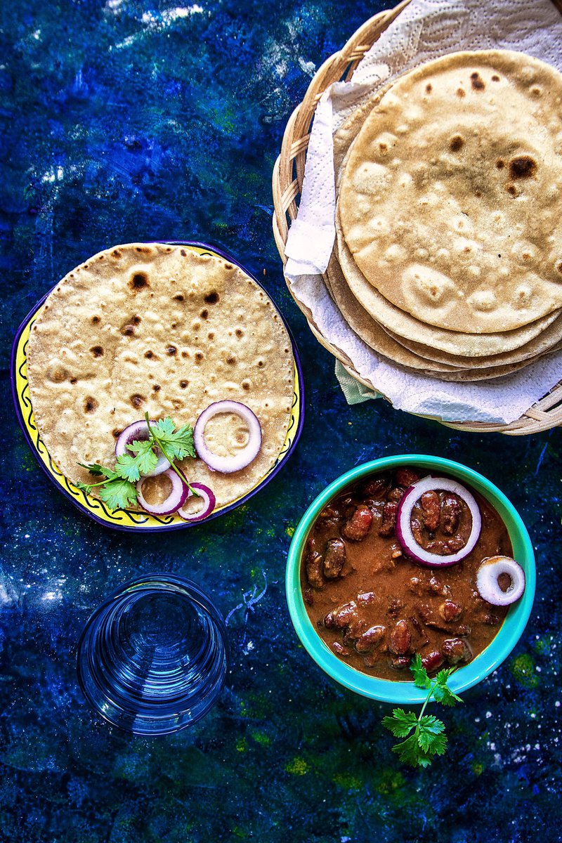 Overhead view of a roti plate, bowl of rajma masala aka kidney bean curry, and glass of water on a blue background