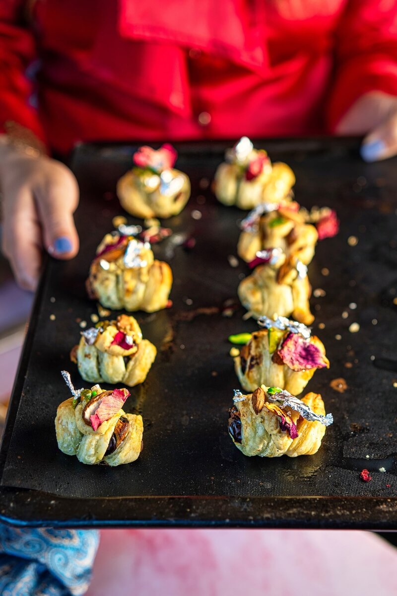 Freshly baked Burfi Stuffed Puff Pastry Date Bites on a baking tray topped with chopped nuts and edible silver leaf