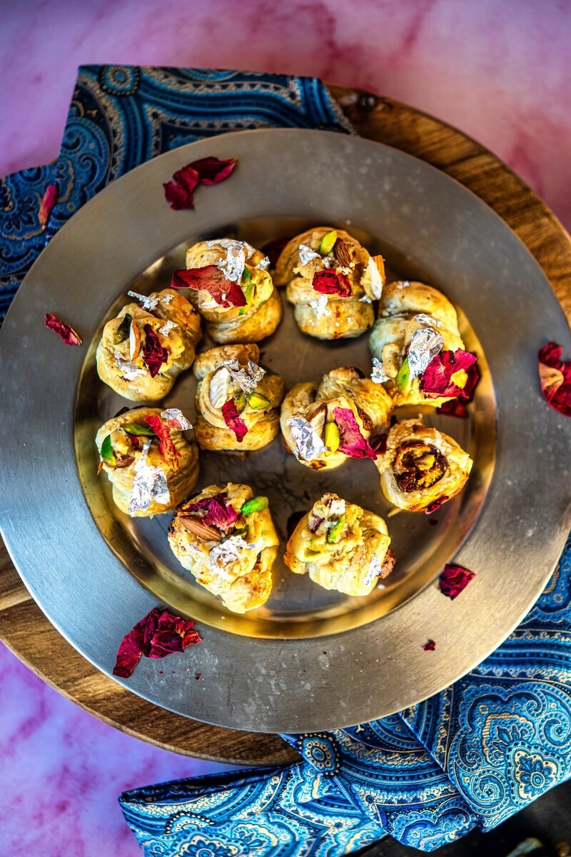 Overhead view of Royal Burfi Date Puff Bites arranged on a silver plate with rose petals and nuts for Diwali celebration