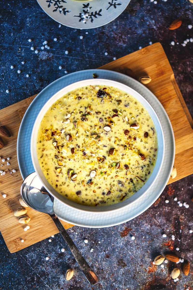 A bowl of sabudana kheer garnished with nuts, placed on a wooden board with a spoon beside it.