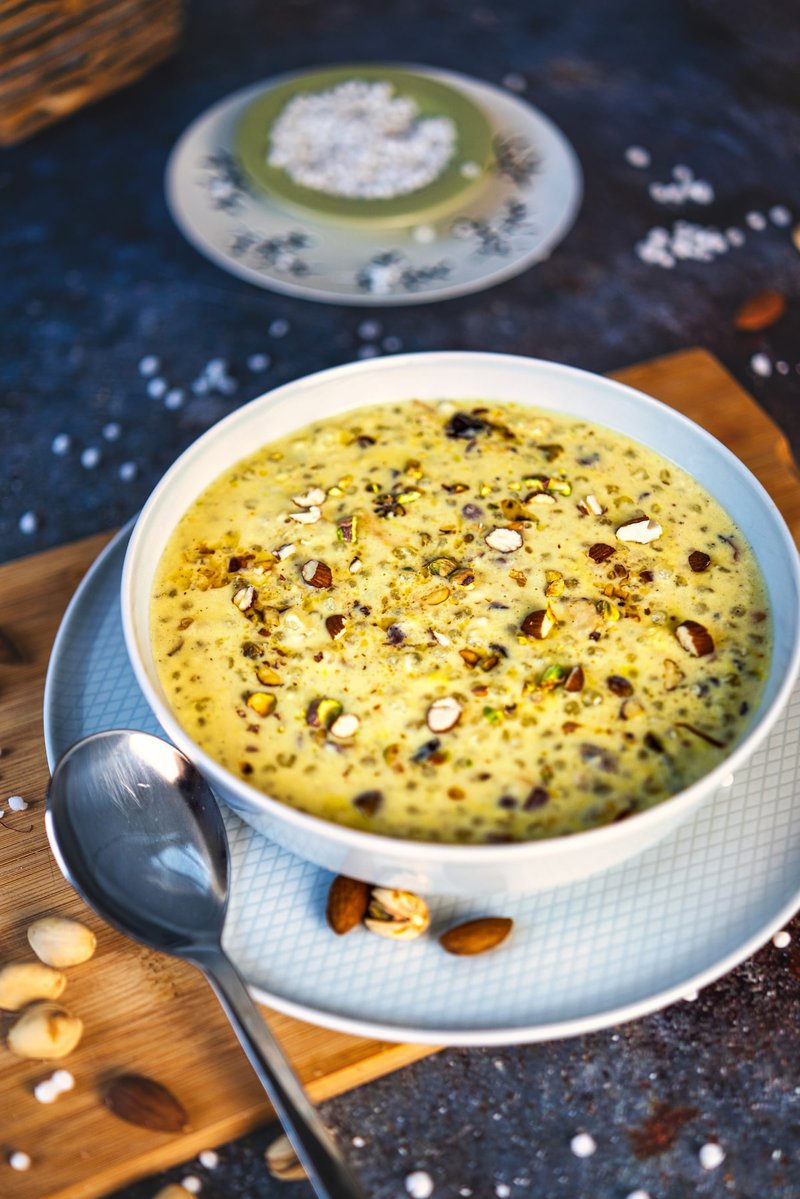 Bowl of creamy sabudana kheer topped with nuts, with a spoon and a plate of sabudana in the background.