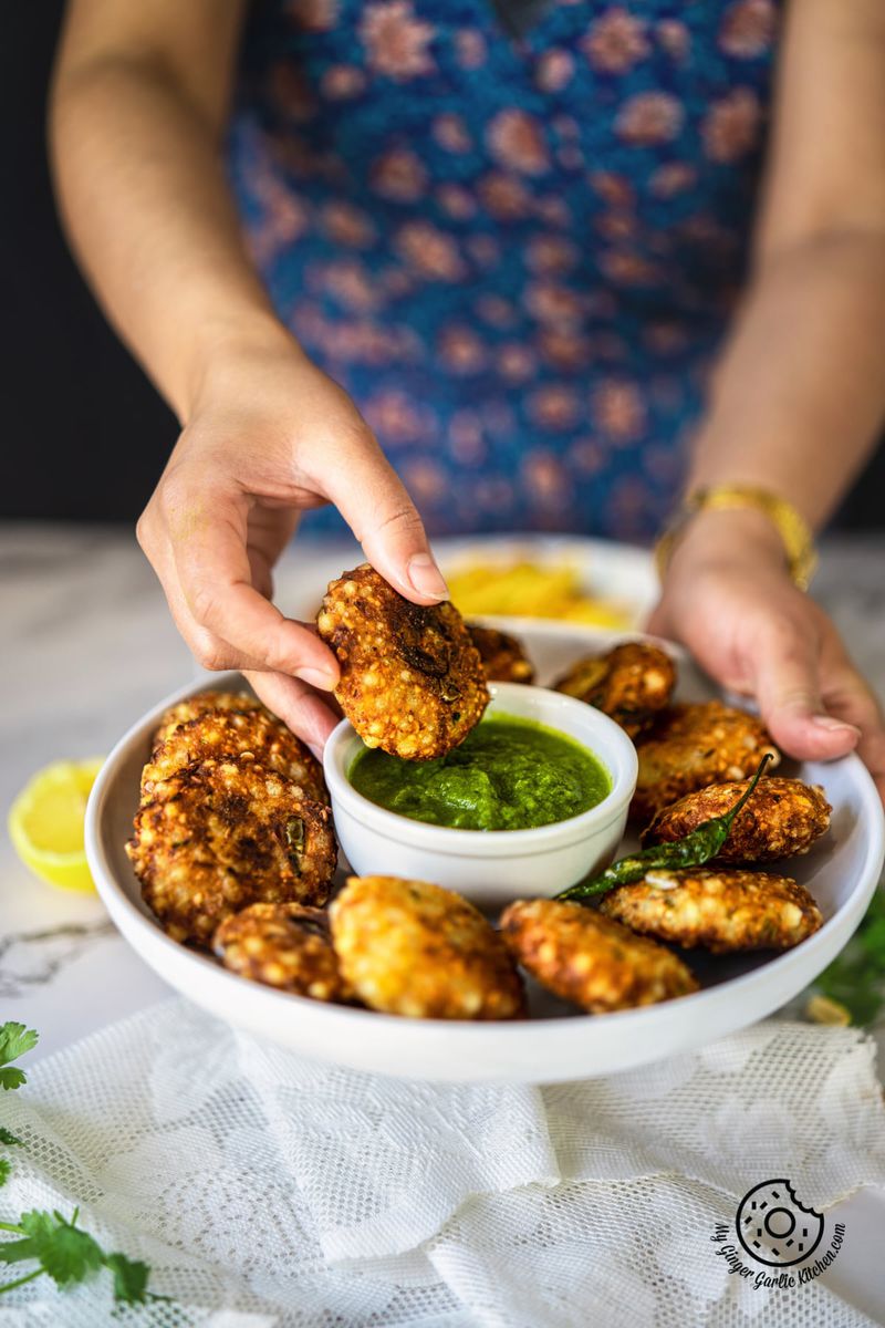 a female holding a sabudana vada over green chutney bowl kept in a white plate with more vadas
