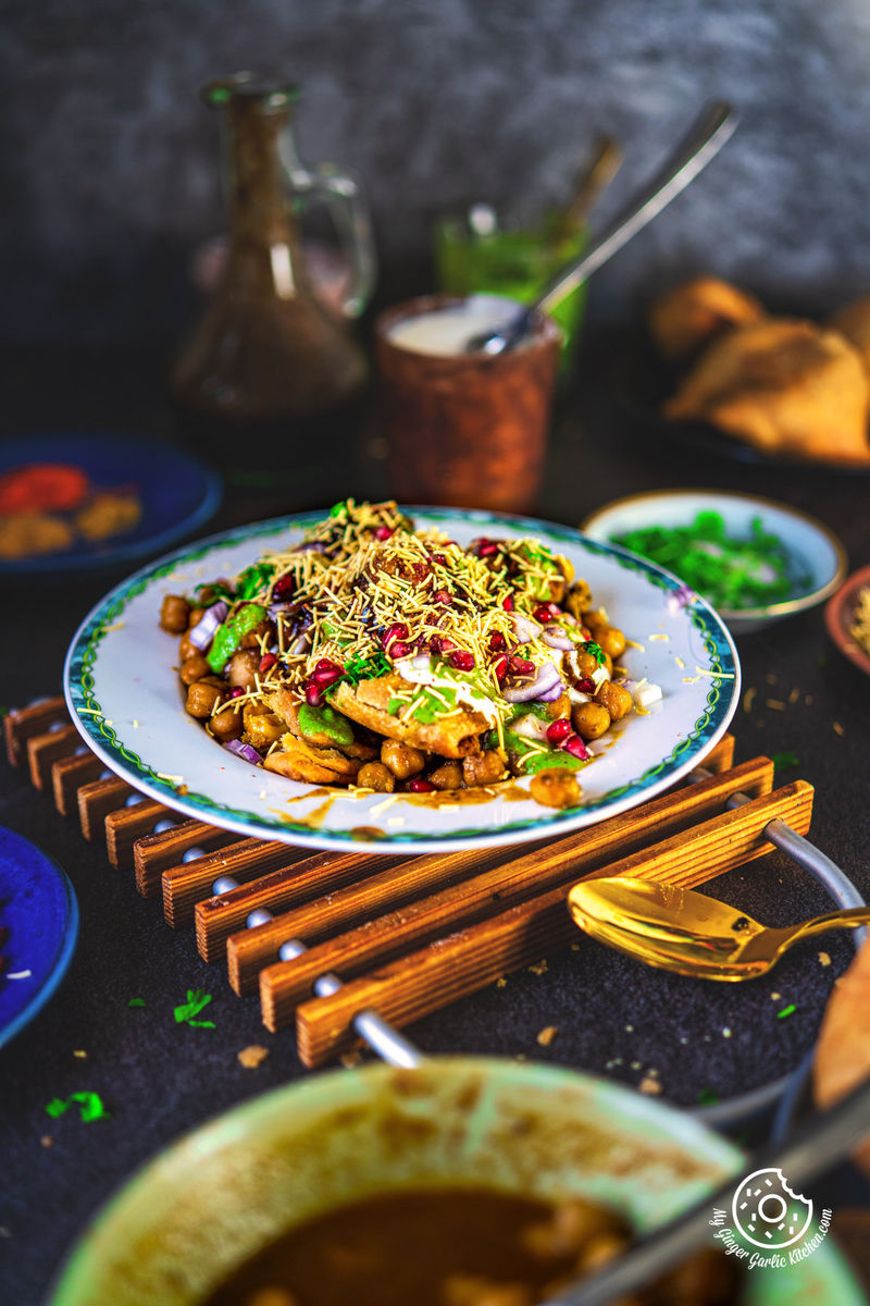 A vibrant dish of samosa chaat garnished with sev, pomegranate seeds, and chopped herbs, presented on a decorative plate, with a backdrop of a rustic jug, a cup of chai, and dimly lit ambiance.
