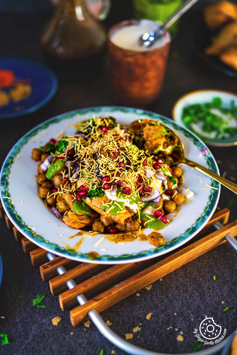 Colorful and appetizing samosa chaat topped with crunchy sev, fresh pomegranate, and diced onions on an ornate plate, accompanied by Indian condiments and a traditional metal cup.