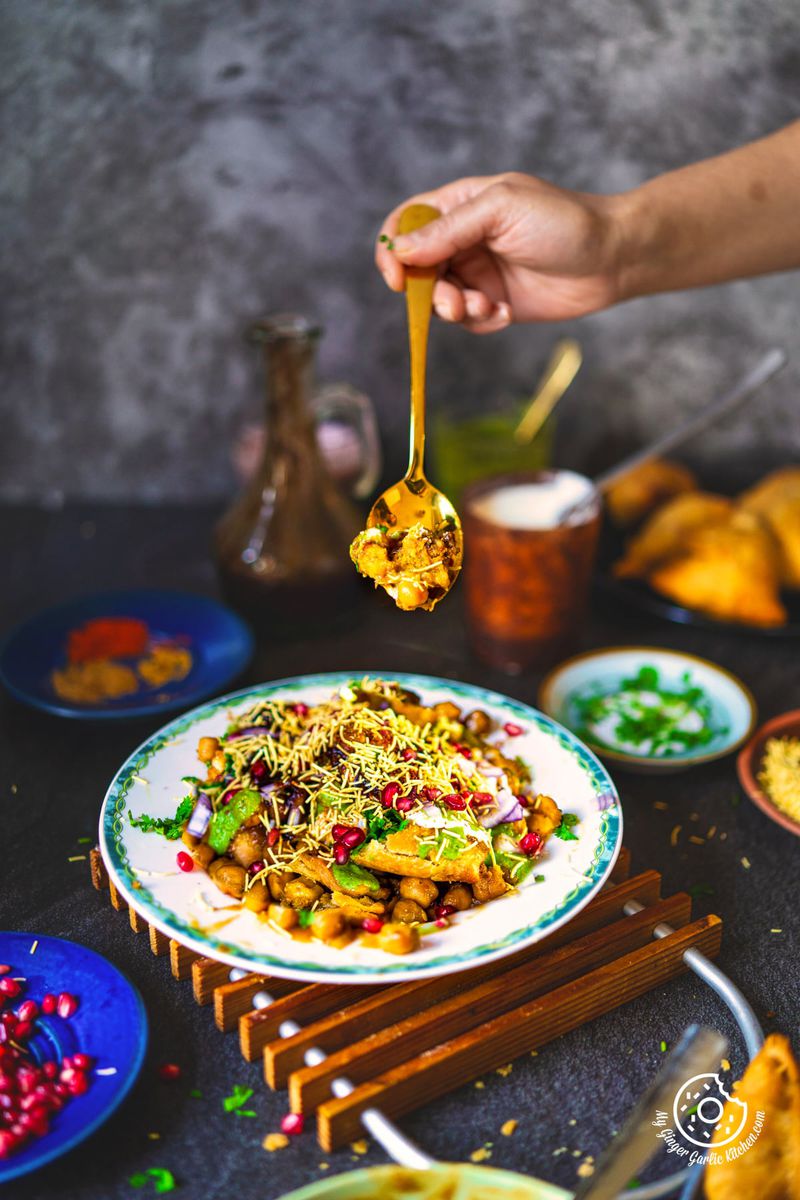 A person's hand holding a golden spoon, scooping up a portion of samosa chaat, showcasing the texture and toppings of this popular Indian street food. There is also a plate full samosa in the background