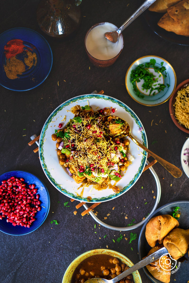 Overhead shot of samosa chaat richly garnished with sev and pomegranate, surrounded by a selection of Indian snacks and dips, embodying a feast for the senses.