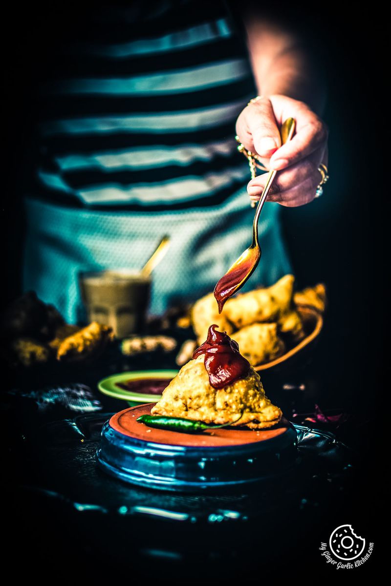 a girl dripping some ketchup with a golden spoon on samosa