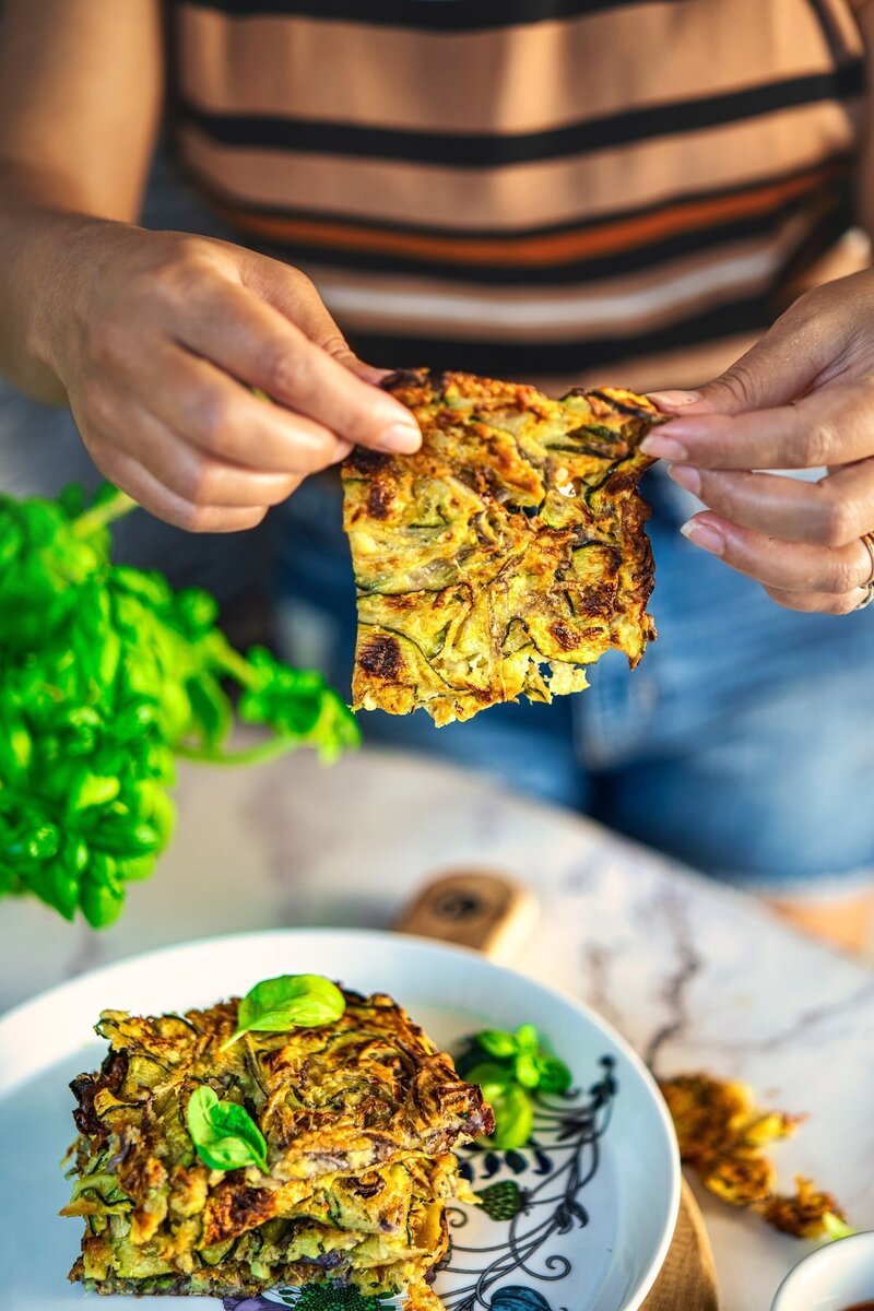 IHand holding thin, crispy Tuscan scarpaccia zucchini tart above plate of savory squares