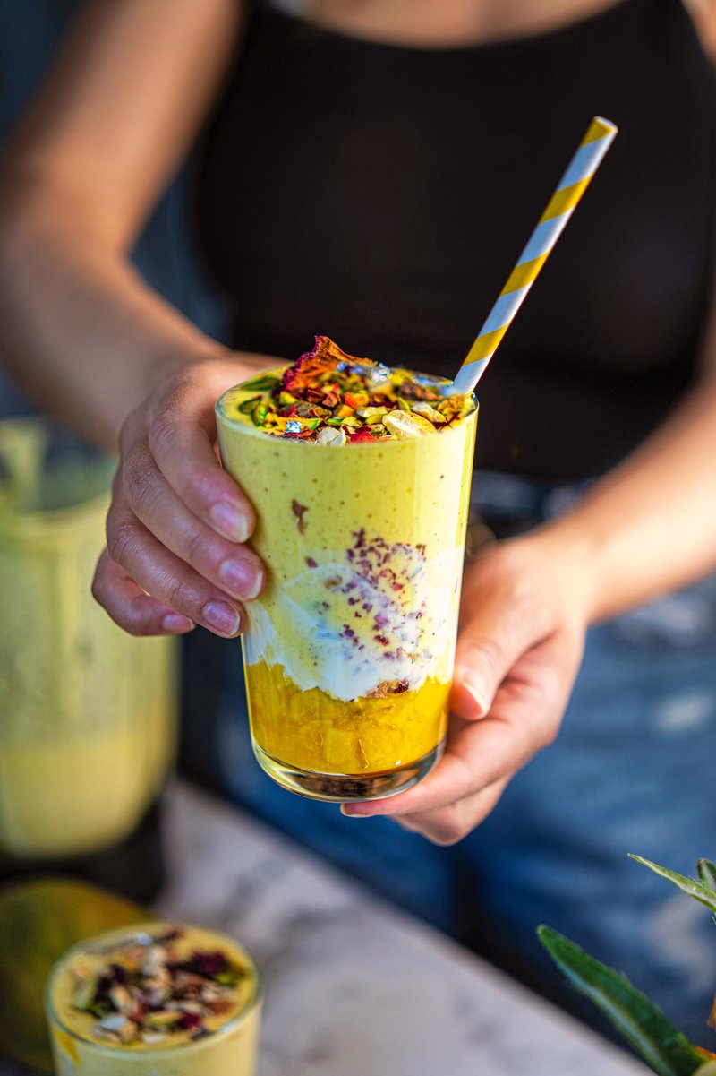 Hand holding a glass of thick and creamy Shahi Mango Lassi with mango chunks, yogurt decoration, nuts, rose petals, saffron, and edible silver leaf.