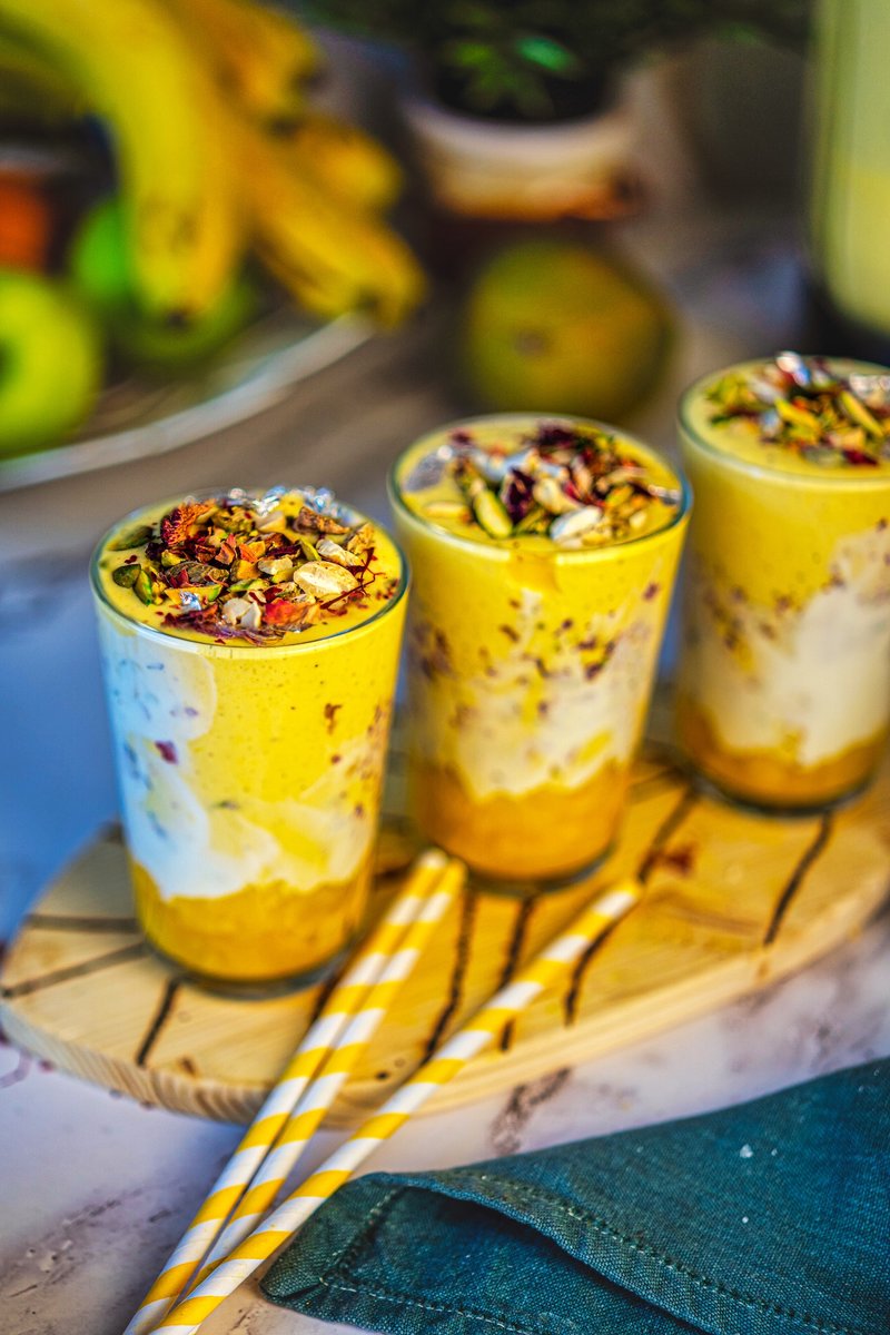 Three glasses of Shahi Mango Lassi topped with chopped pistachios, almonds, rose petals, saffron, and edible silver leaf, served in yogurt-lined glasses with a layer of muddled mangoes at the bottom.