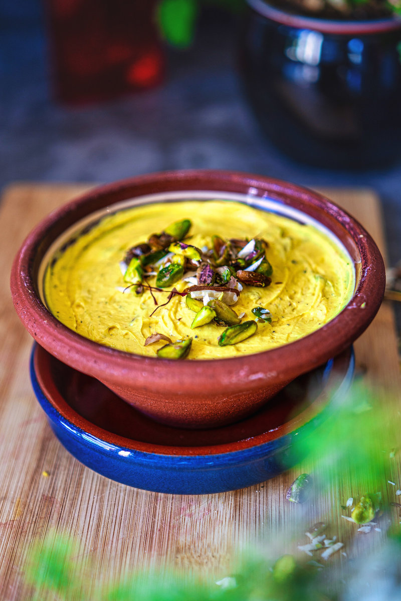 Shrikhand in a rustic brown clay bowl garnished with chopped nuts, placed on a wooden table with blurred greenery in the background.