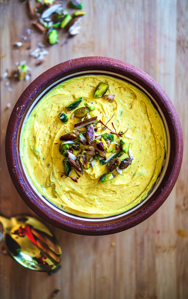 Top view of shrikhand in a brown clay bowl with a garnish of pistachios and almonds on a wooden table.