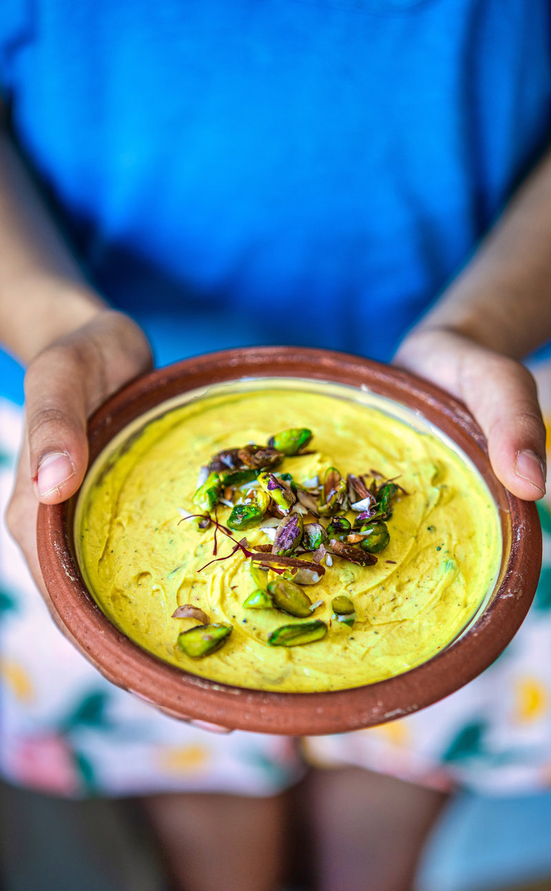 Person holding a clay bowl of shrikhand garnished with nuts, showcasing the dessert at an outdoor setting.